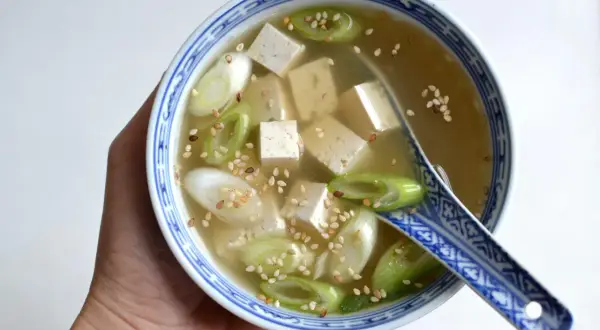 Miseczka z bulionem miso, kostkami tofu, zieloną cebulką i sezamem, trzymana w dłoni.