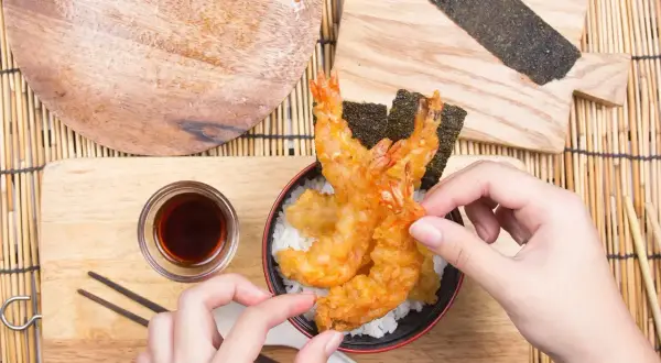 Ręce sięgają po chrupiące krewetki w panierce tempura, ułożone na ryżu z nori.