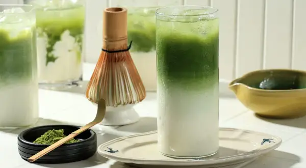 Orzeźwiające matcha latte z lodem, idealne dla mam karmiących piersią. Obok bambusowa miotełka i łyżeczka do przygotowania napoju.
