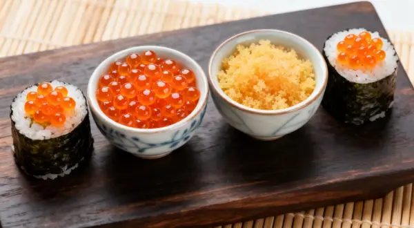 Dwa sushi z ikrą łososia i miseczki z ikrą masago i ikrą łososia na drewnianej desce.