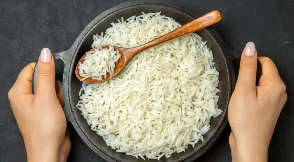 Dwie dłonie trzymają okrągłą patelnię z parującym ryżem basmati. Drewniana łyżka zanurzona w ryżu.