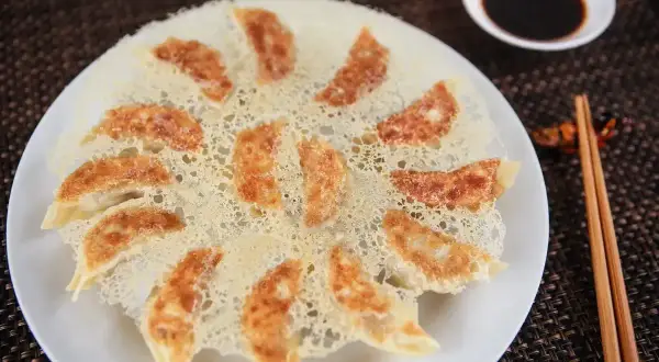 Chrupiące pierożki gyoza ułożone na talerzu, z chrupiącą panierką z ciasta. Obok miseczka sosu sojowego i pałeczki.