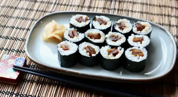 Tykwa sushi, czyli rolki z ryżem i ciemnym nadzieniem, ułożone na talerzu obok imbiru i pałeczek.