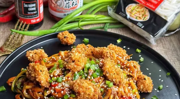 Pyszny udon z kurczakiem i warzywami, posypany sezamem i szczypiorkiem. Obok sos sojowy i opakowanie makaronu udon.