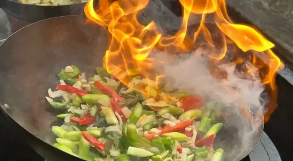 Płomienie otaczają warzywa w woku, tworząc apetyczny stir fry. W tle widać drugie danie.