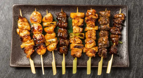 Różnorodne szaszłyki yakitori z kurczakiem i warzywami, polane słodkim sosem, ułożone na ciemnej tacy.