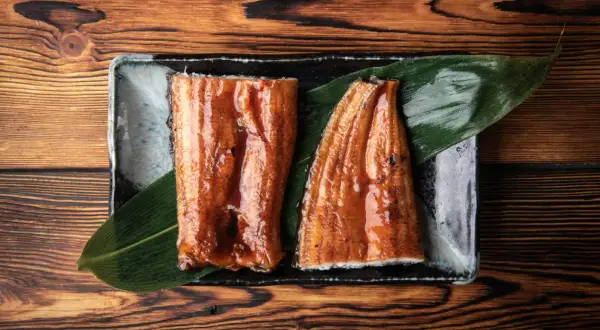 Dwa kawałki lśniącego, glazurowanego unagi na liściu, podane na kamiennej tacy na drewnianym tle.