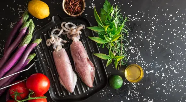 Dwa surowe kalmary na grillu, obok pomidorów, bakłażanów, cytryny, limonki i ziół. Idealne składniki, by dowiedzieć się, jak jeść kalmary.