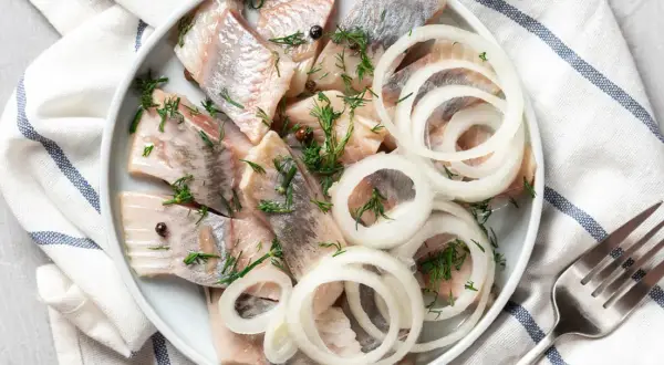 Pyszny śledź a'la matjas na talerzu, udekorowany krążkami cebuli i koperkiem. Gotowy do podania z widelcem obok.