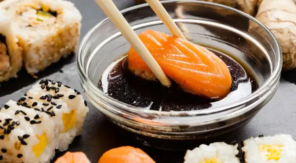 Paluszki zanurzają kawałek łososia w sosie sojowym bezglutenowym. Obok sushi i imbir.