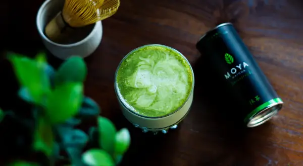 Szklanka zielonego matcha z pianką, obok puszka MOYA Matcha i bambusowa miotełka.