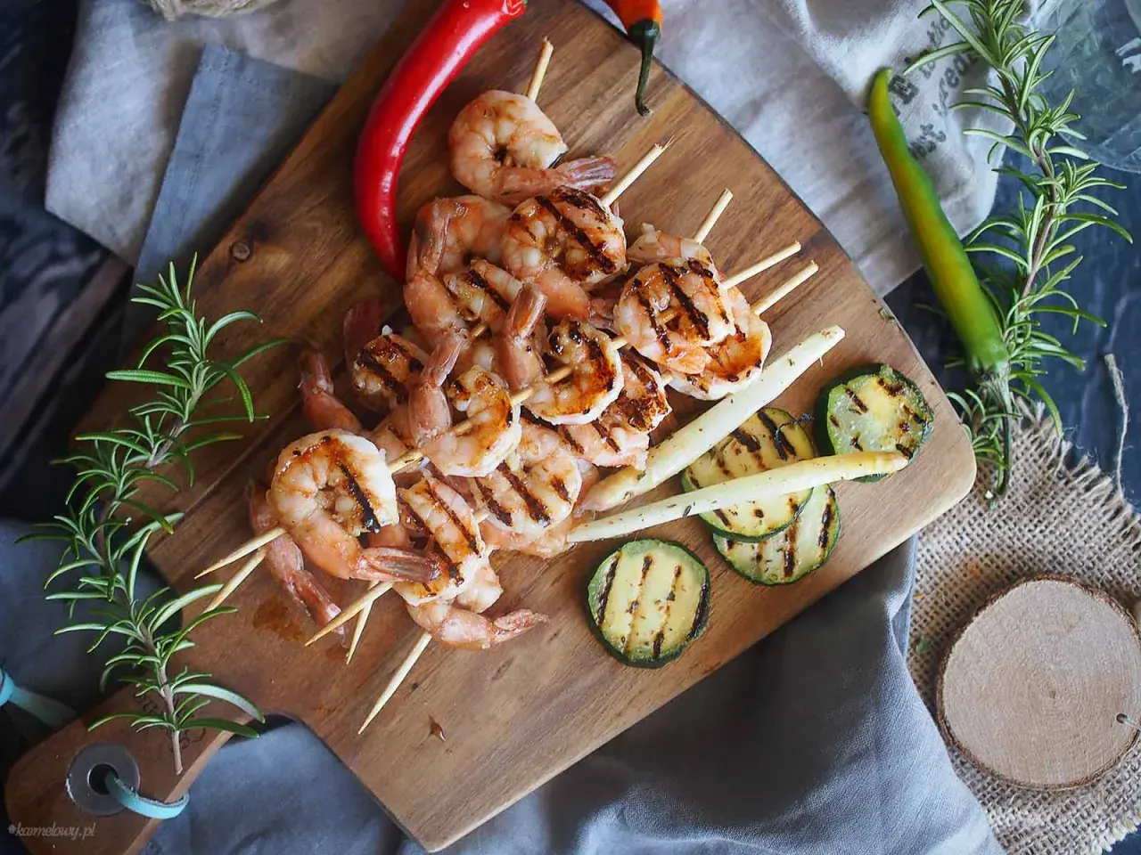Grillowane krewetki po angielsku na szpadkach, podane z grillowaną cukinią, szparagami i papryczkami chili na drewnianej desce.