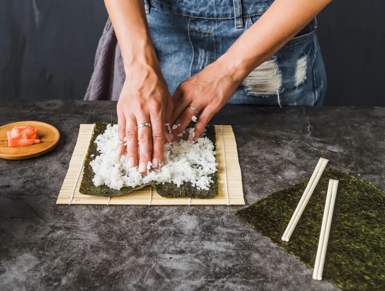 Dłonie rozprowadzają ryż haruka na nori, przygotowując się do zwinięcia sushi. Obok leżą pałeczki i marynowany imbir.