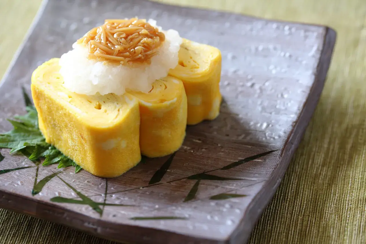 Tamago co to? Japoński omlet tamagoyaki, zwinięty w rolki, podany z tartą rzodkwią i grzybami enoki na talerzu z bambusowym wzorem.