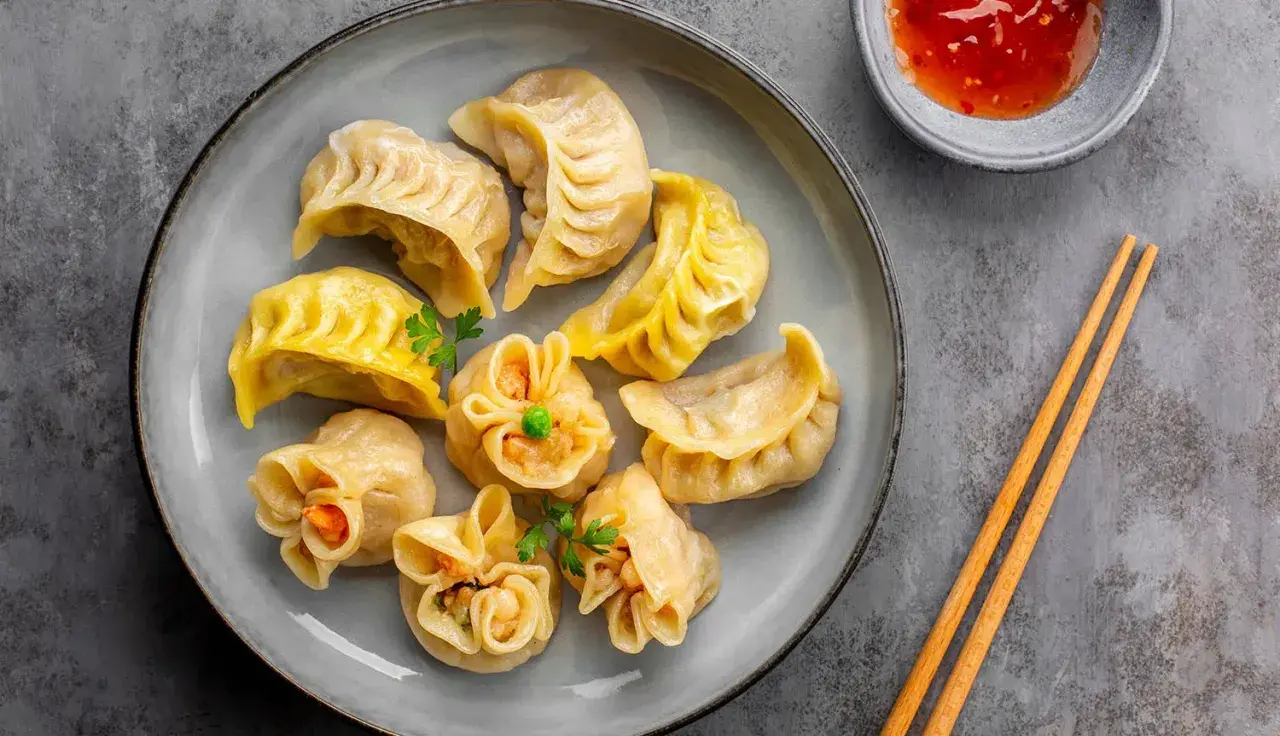 Talerz pełen różnorodnych pierożków dim sum, ozdobionych natką pietruszki, z miseczką sosu i pałeczkami.