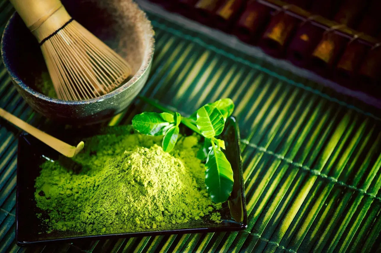 Zielony proszek matcha na czarnym talerzyku, bambusowa miotełka i liście.