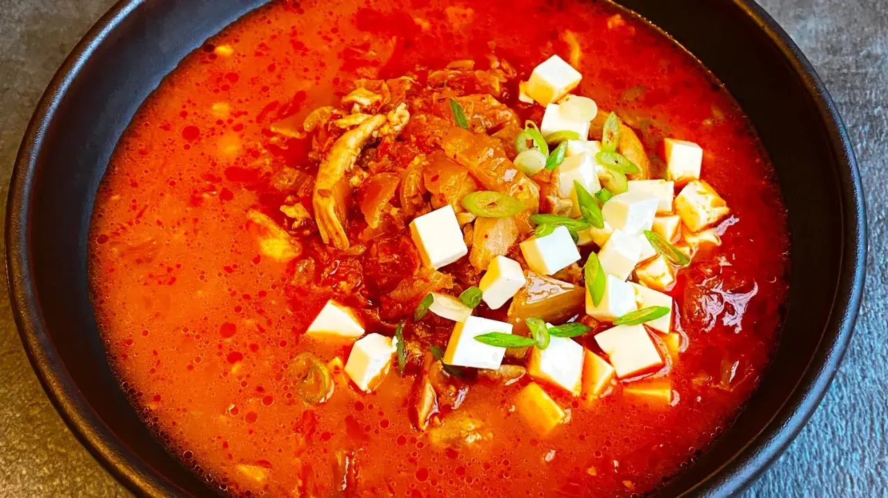 Pikantna zupa kimchi z kurczakiem i kostkami tofu, udekorowana szczypiorkiem.