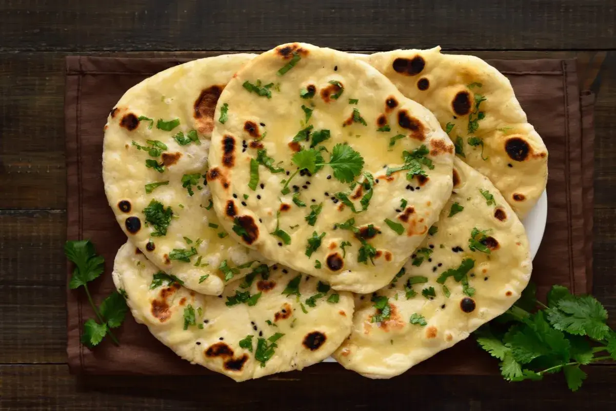 Stos chlebków naan, posypanych kolendrą i czarnuszką, z widocznymi pęcherzykami powietrza. Idealne do curry, ale warto sprawdzić chlebek naan kcal.