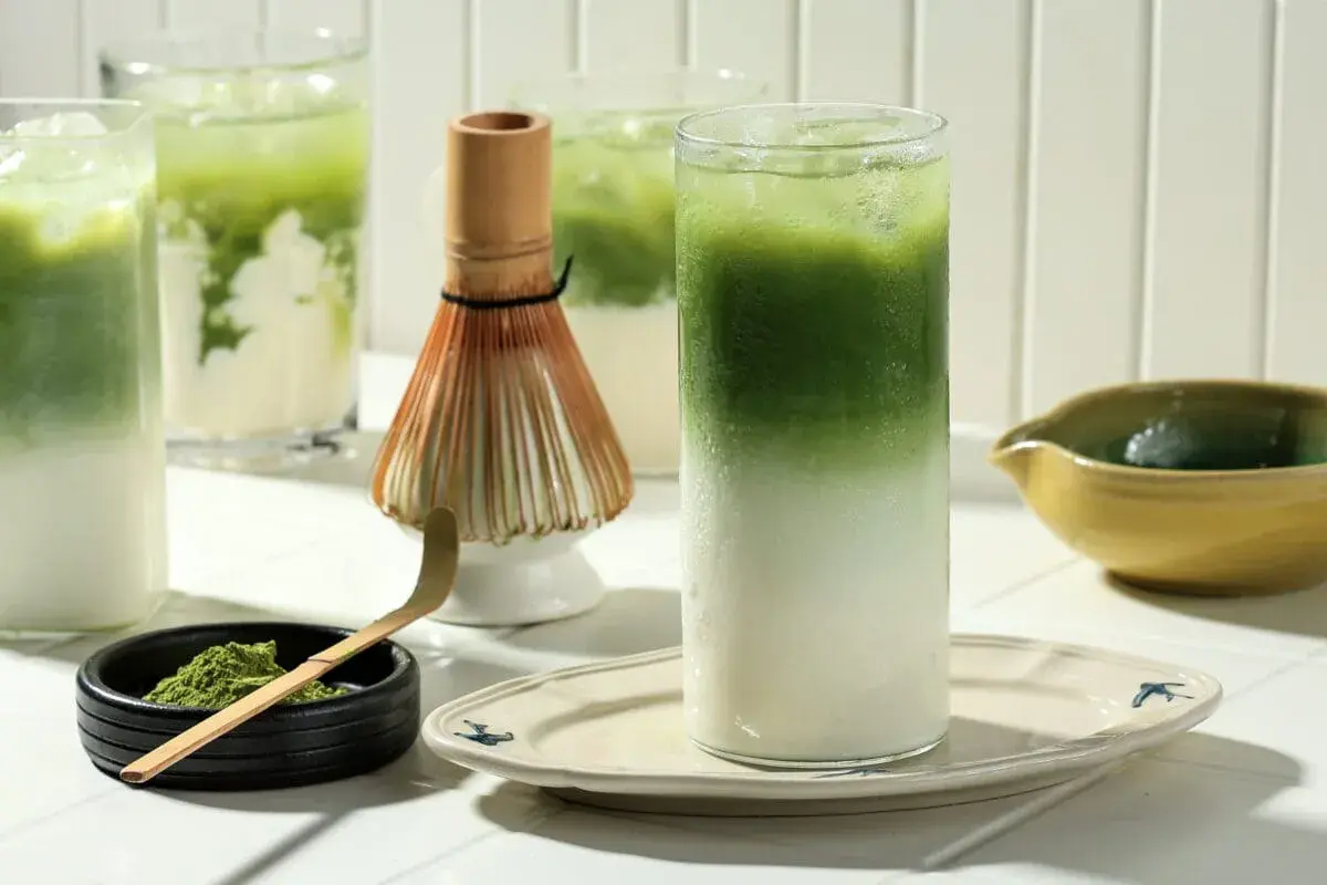 Orzeźwiające matcha latte z lodem, idealne dla mam karmiących piersią. Obok bambusowa miotełka i łyżeczka do przygotowania napoju.
