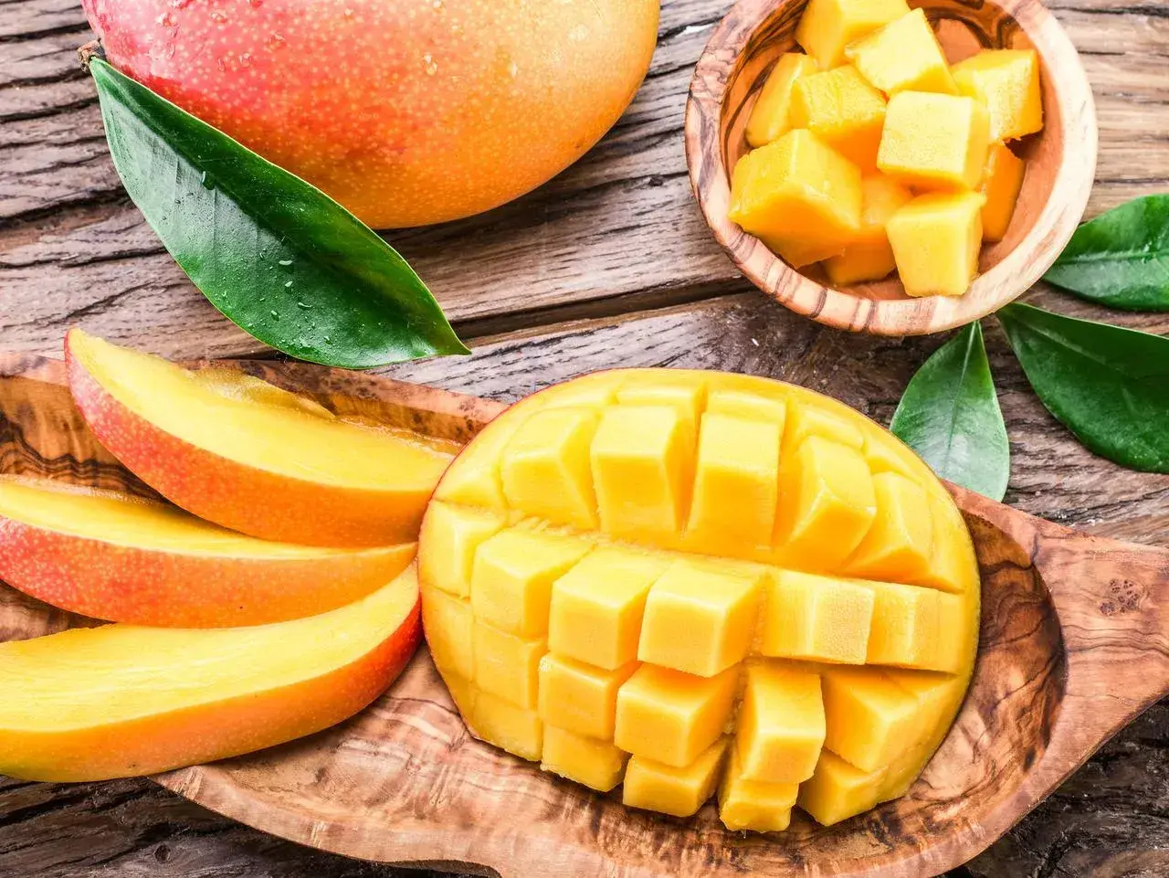 Soczyste, pokrojone w kostkę i plastry mango, które pokazuje, co to jest mango. Całe, dojrzałe owoce i liście na drewnianym tle.