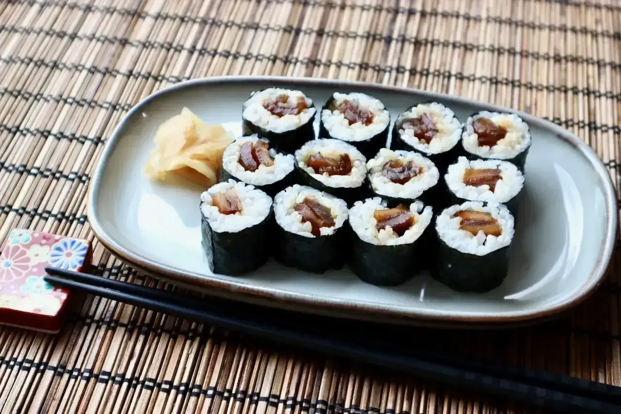 Tykwa sushi, czyli rolki z ryżem i ciemnym nadzieniem, ułożone na talerzu obok imbiru i pałeczek.