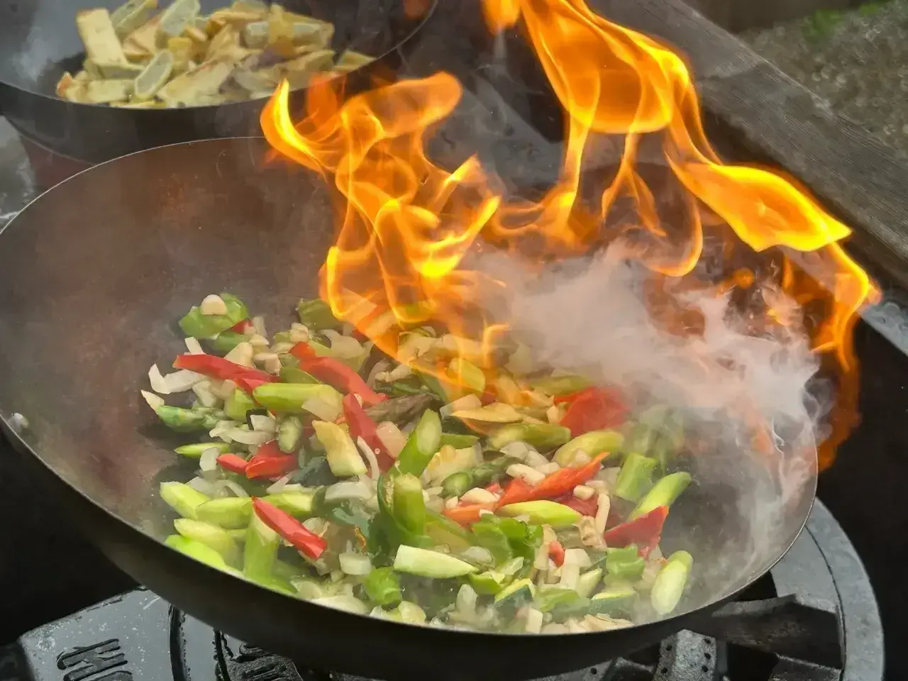 Płomienie otaczają warzywa w woku, tworząc apetyczny stir fry. W tle widać drugie danie.