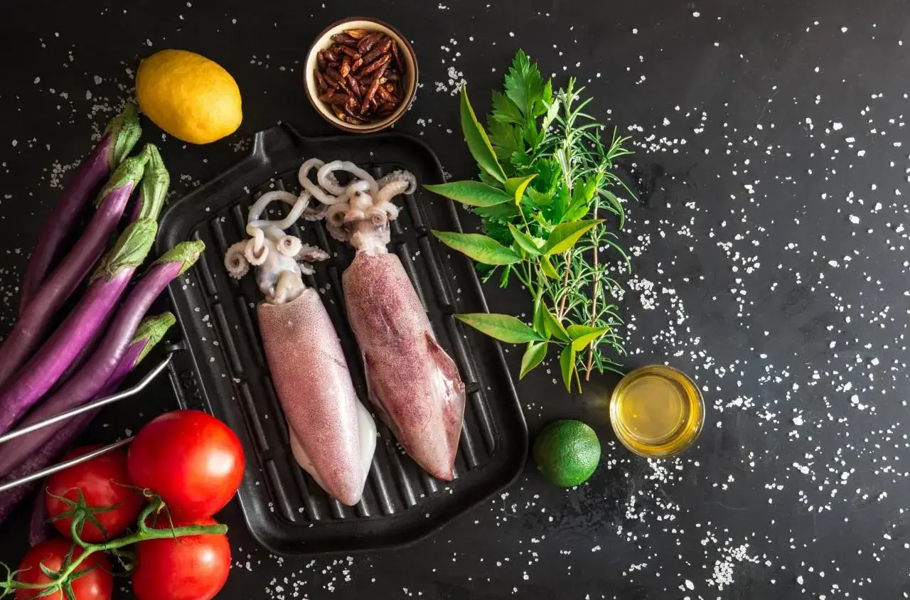 Dwa surowe kalmary na grillu, obok pomidorów, bakłażanów, cytryny, limonki i ziół. Idealne składniki, by dowiedzieć się, jak jeść kalmary.