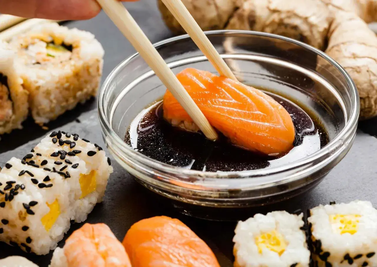 Paluszki zanurzają kawałek łososia w sosie sojowym bezglutenowym. Obok sushi i imbir.