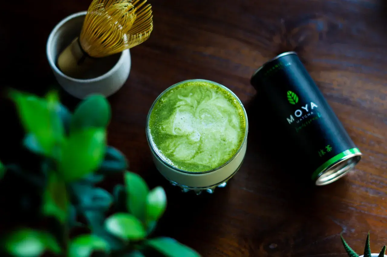 Szklanka zielonego matcha z pianką, obok puszka MOYA Matcha i bambusowa miotełka.