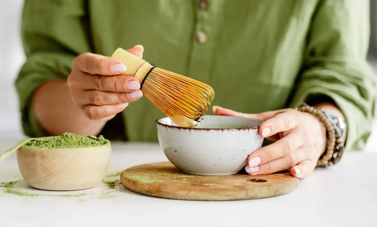 Przygotowanie najlepszej zielonej herbaty matcha. W rękach bambusowa miotełka do spieniania, obok miseczka z zielonym proszkiem.
