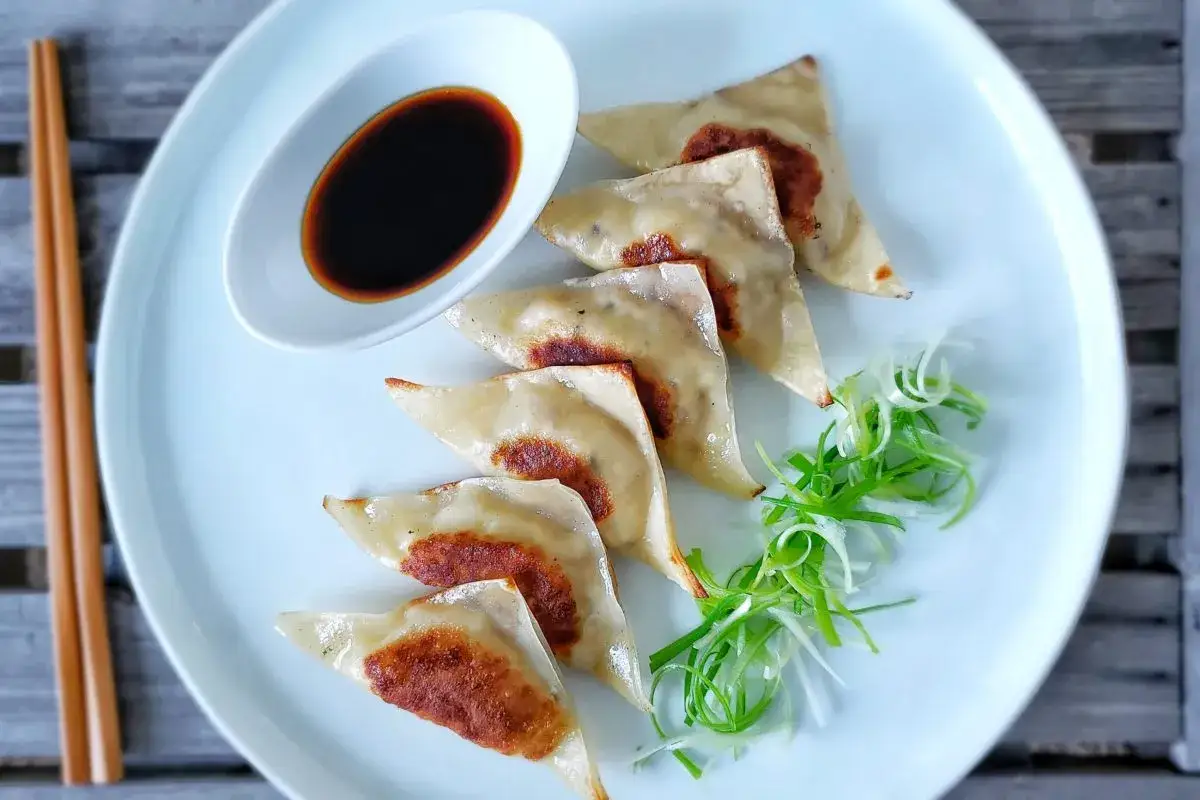 Siedem złocistych pierożków gyoza ułożonych na talerzu, obok sosu sojowego i posiekanej dymki.