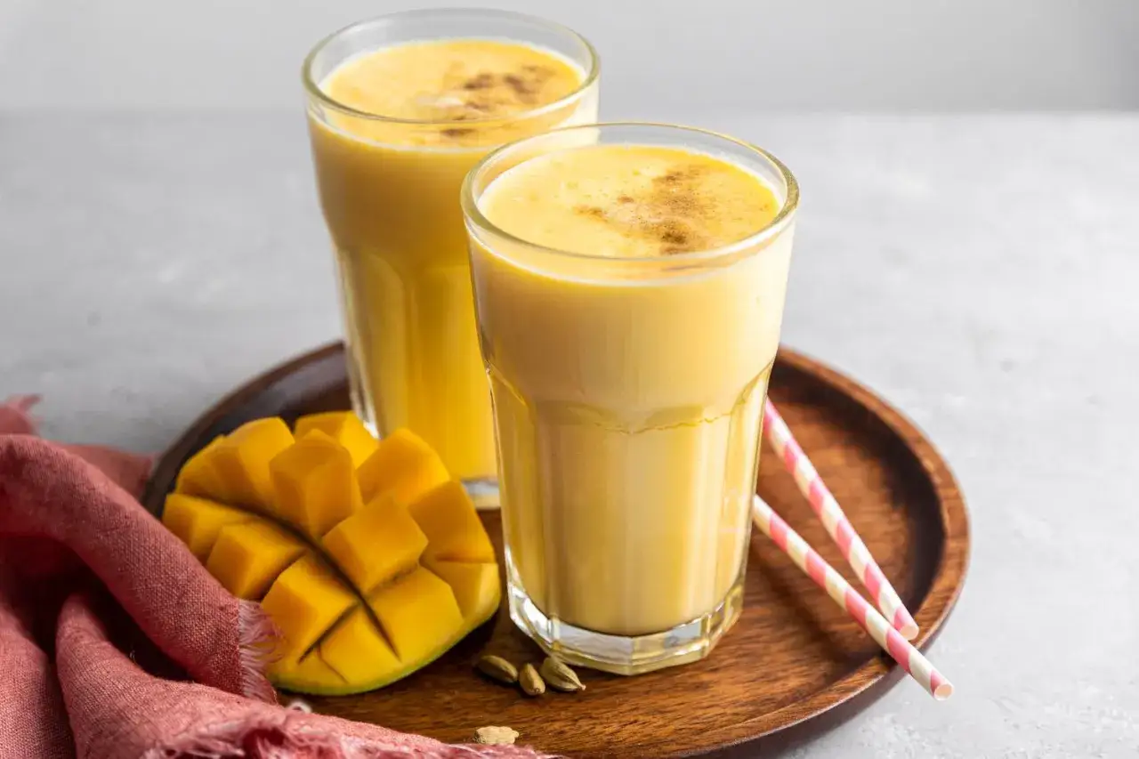 Dwa szklanki kremowego jogurtu z mango, ozdobione kardamonem, obok świeżego owocu i różowych słomek.