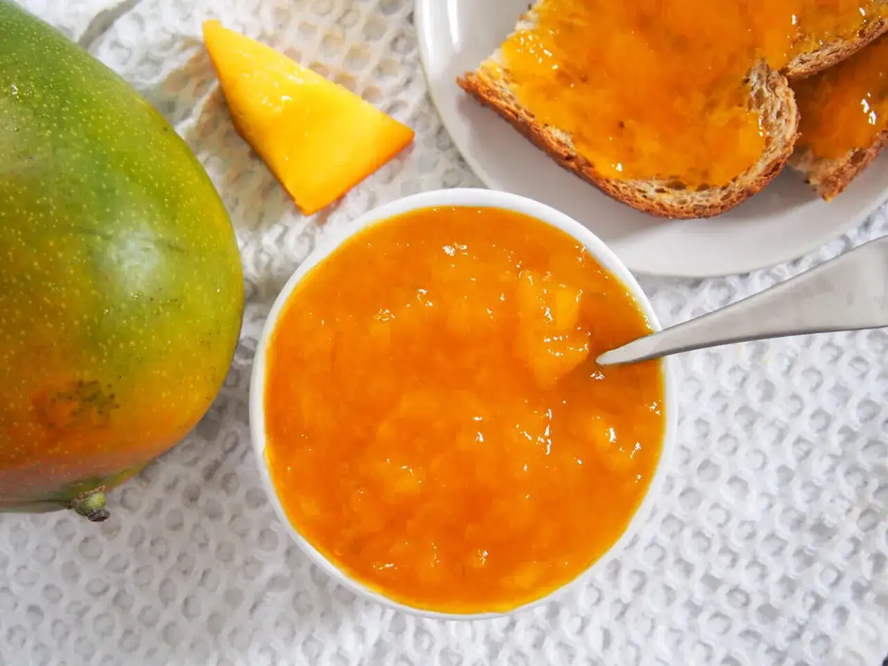 Słoiczek z dżemem z mango, obok świeży owoc i kromki chleba z dżemem. Codzienne jedzenie mango to pyszny początek dnia.
