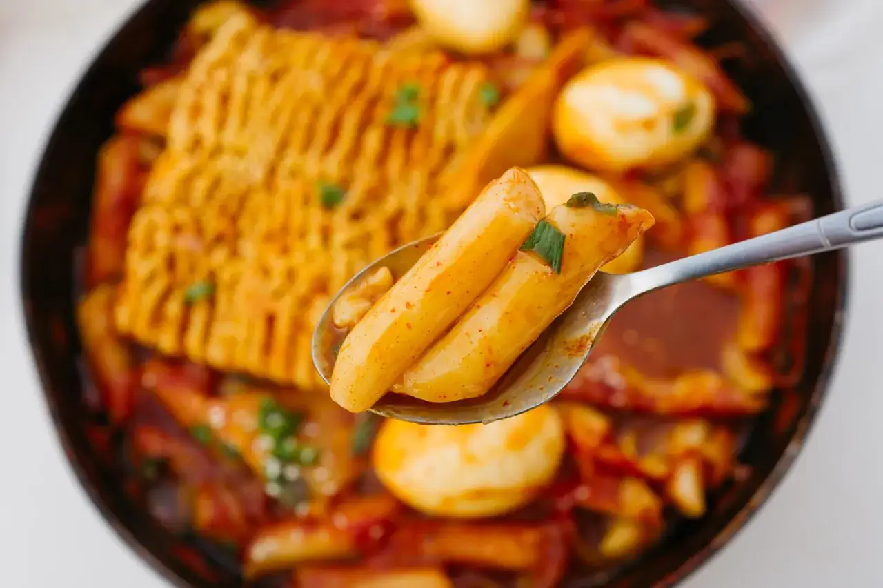 Łyżka z tteokbokki w sosie, obok jajka i placka ryżowego. Pyszne danie, ale warto sprawdzić tteokbokki kcal przed spożyciem.