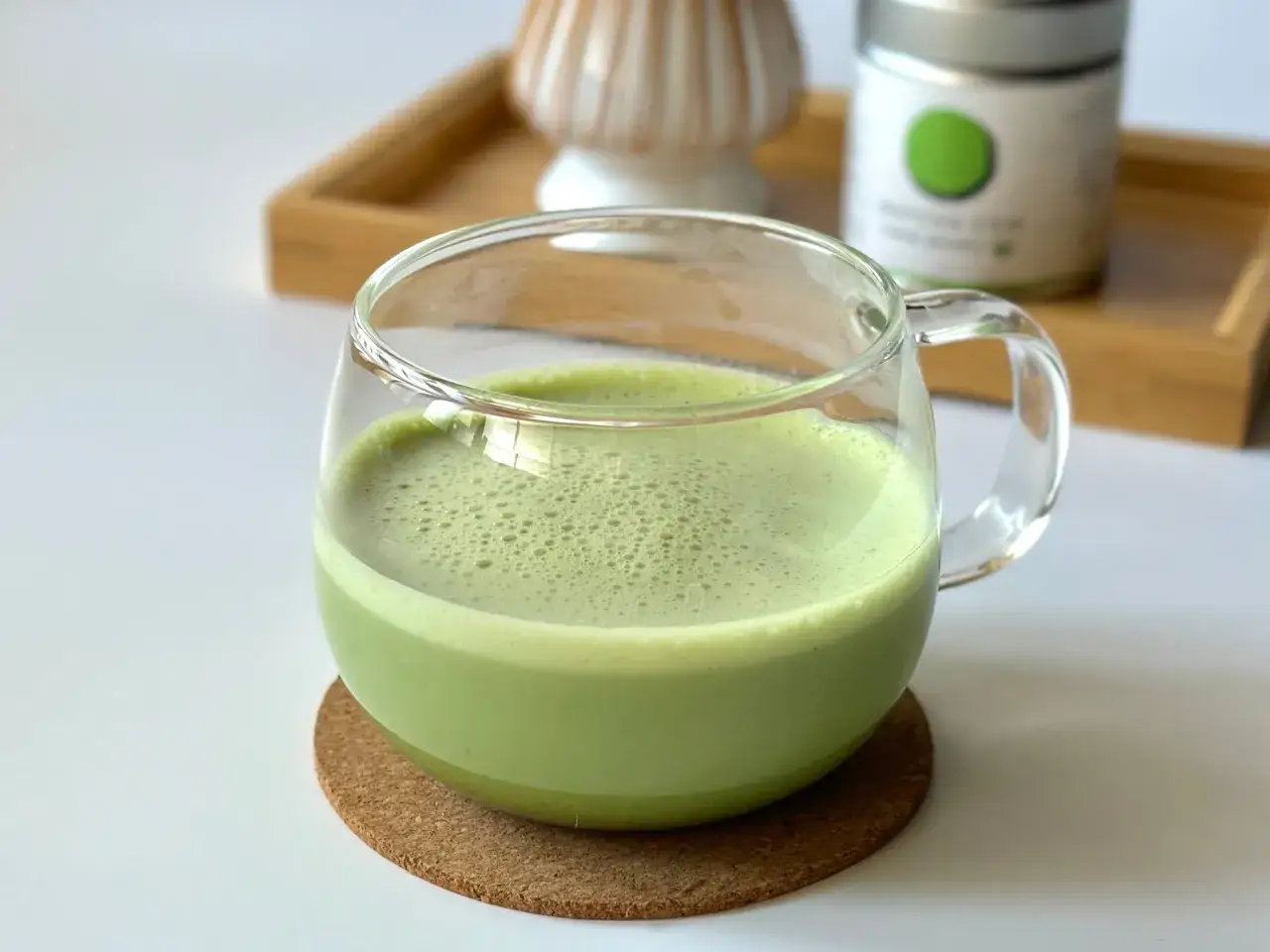 Zielone matcha latte w szklance, idealne dla mam karmiących piersią. Na drewnianej tacce stoi puszka z matchą i bambusowa miotełka.