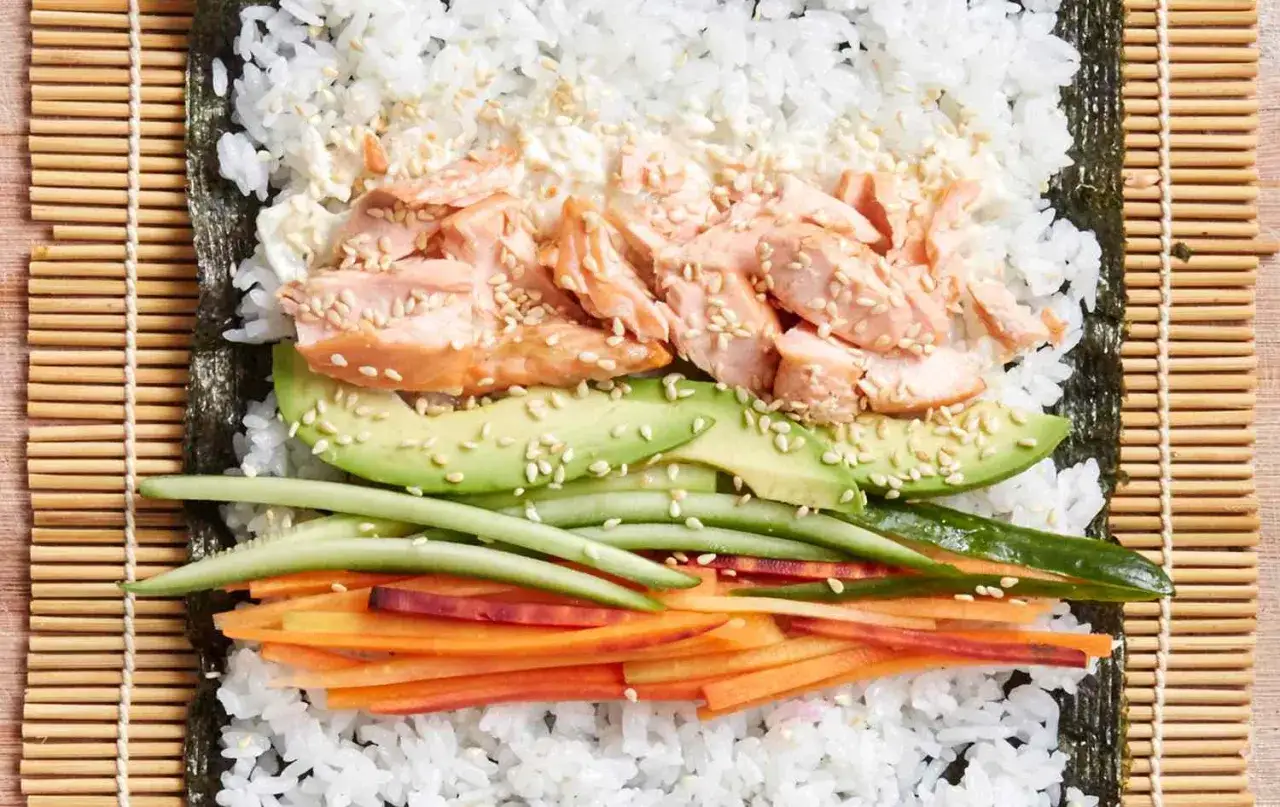 Przygotowanie sushi z łososiem: ryż, nori, awokado, ogórek i marchewka czekają na zwinięcie.
