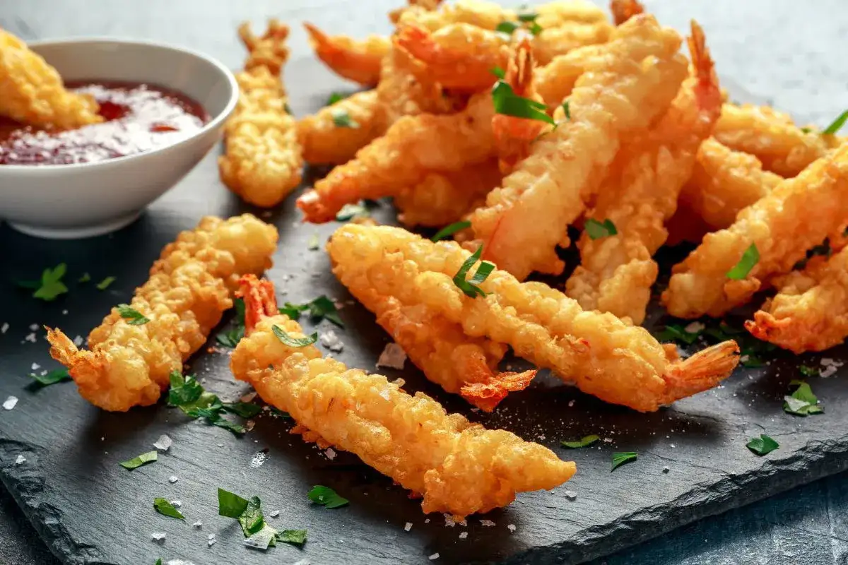 Chrupiące krewetki w panierce tempura, ułożone na ciemnej desce, posypane zieloną pietruszką. Obok miseczka z sosem.