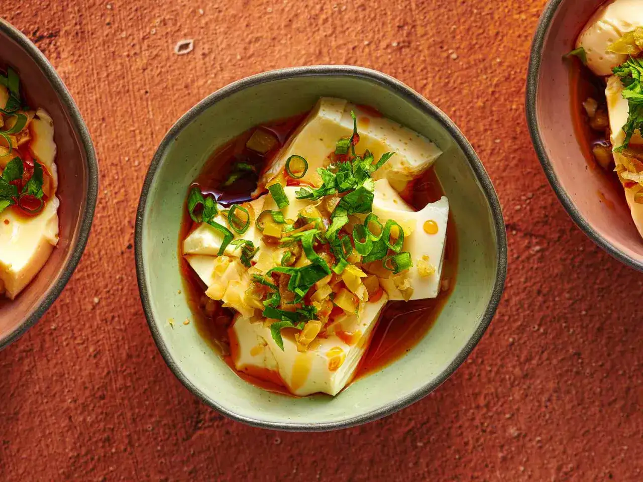 Miękkie tofu w miseczce, polane sosem i posypane szczypiorkiem i innymi dodatkami.