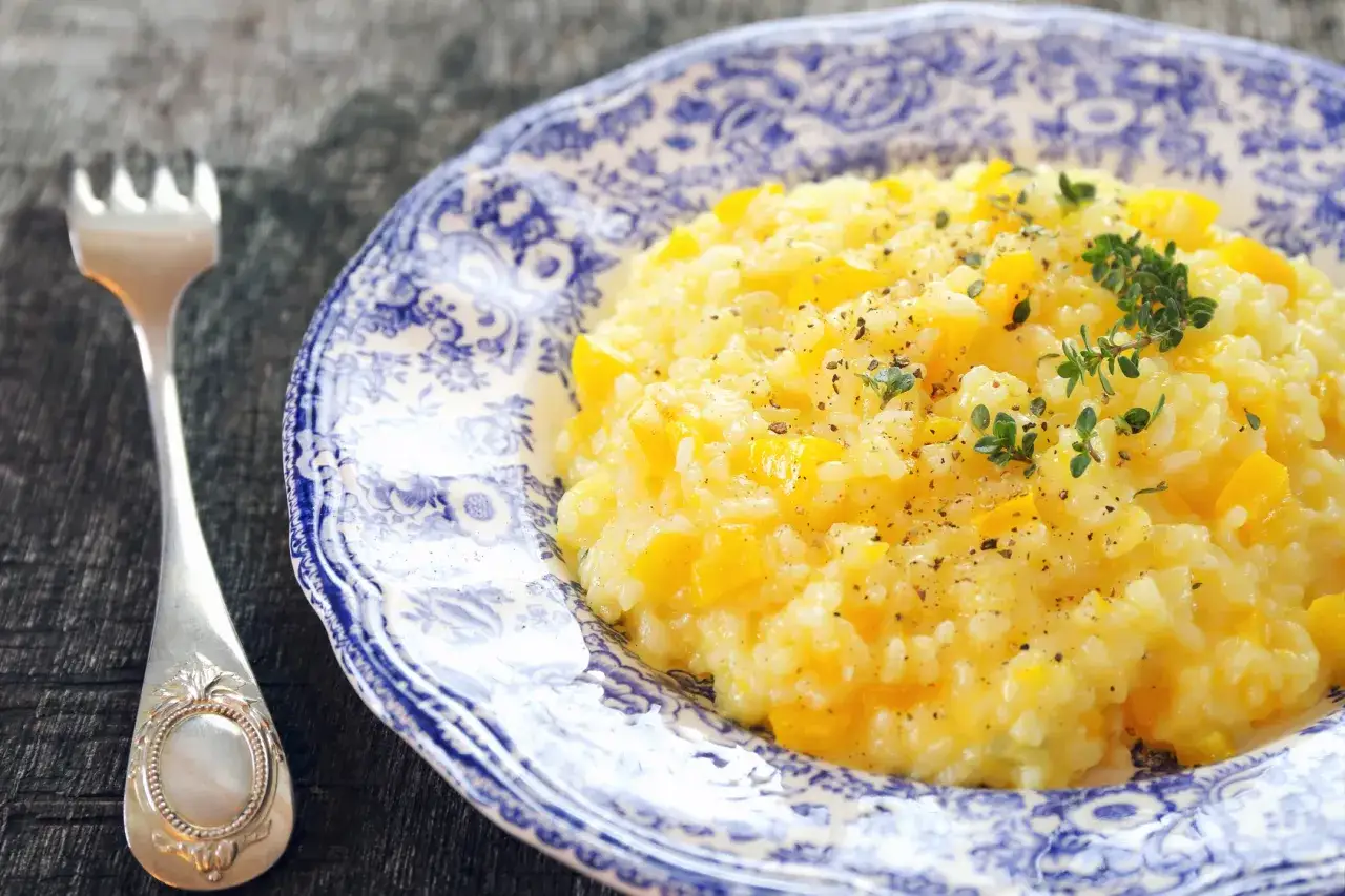 Pyszny, kremowy ryż do risotto z kawałkami dyni, posypany świeżym tymiankiem i pieprzem, na ozdobnym talerzu. Obok leży srebrny widelec.