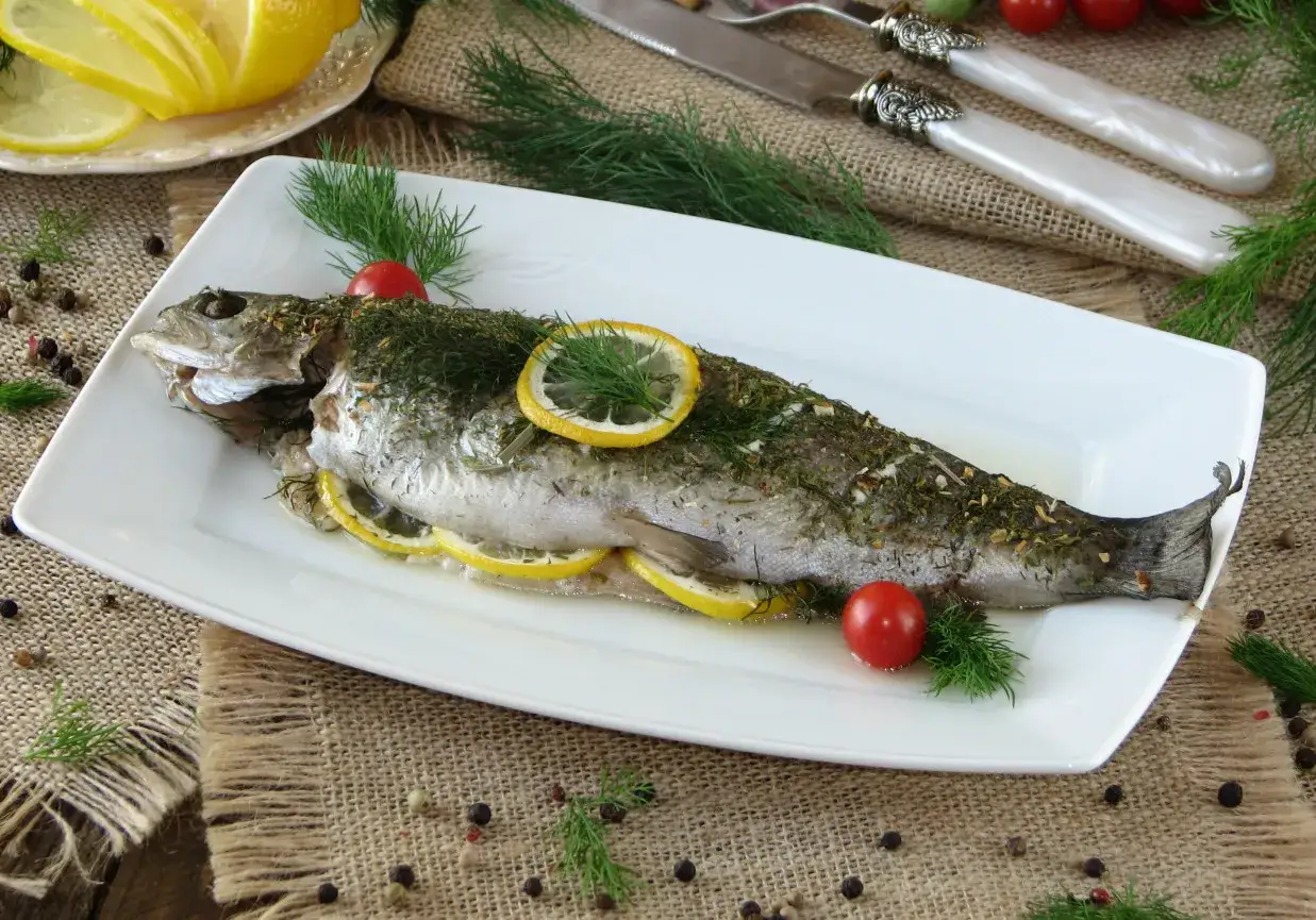 Pstrąg z grilla, pieczony z cytryną i ziołami, podany na białym talerzu z pomidorkami koktajlowymi i koperkiem.