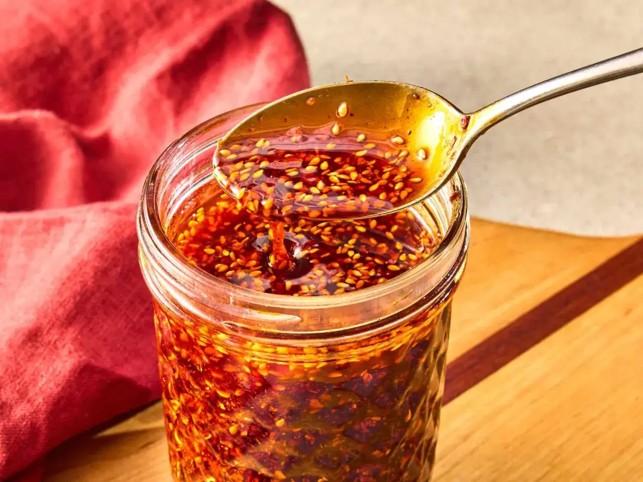 Słoik pełen aromatycznego chili w oleju, z którego łyżka unosi złocisty płyn z nasionami sezamu.