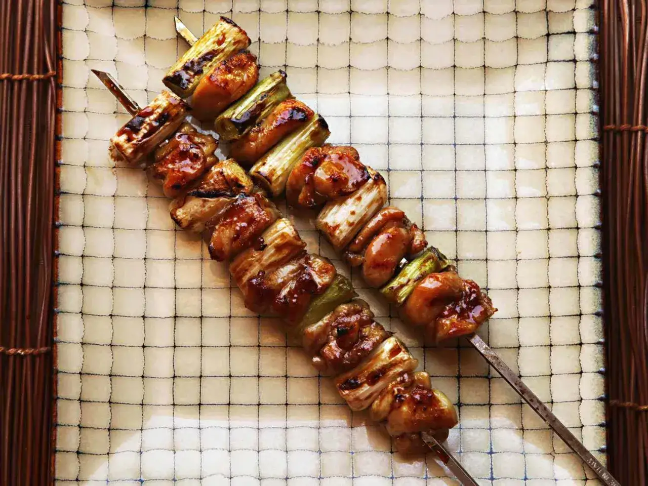 Dwa apetyczne szaszłyki yakitori z kurczakiem i zieloną cebulką, polane słodkim sosem, leżą na talerzu z kratką.