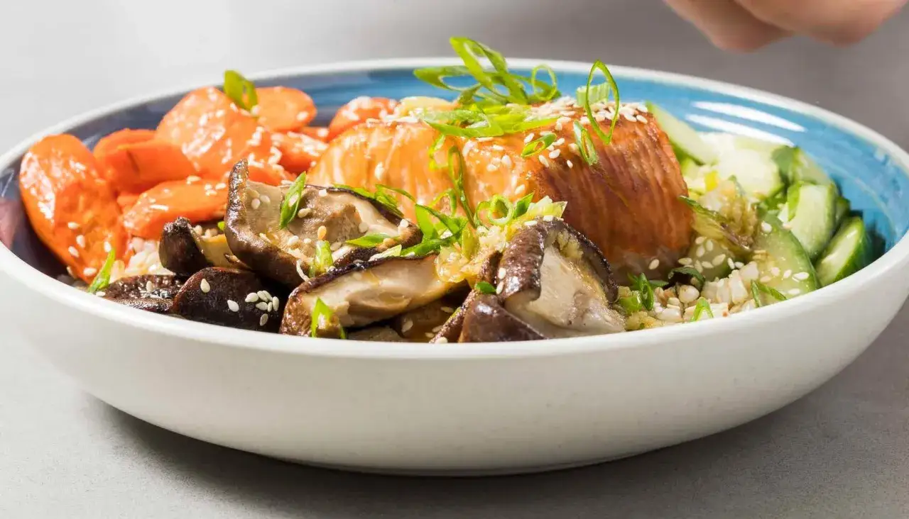 Pyszny bowl z ryzem, łososiem, pieczarkami shiitake, marchewką i ogórkiem, posypany sezamem i szczypiorkiem.