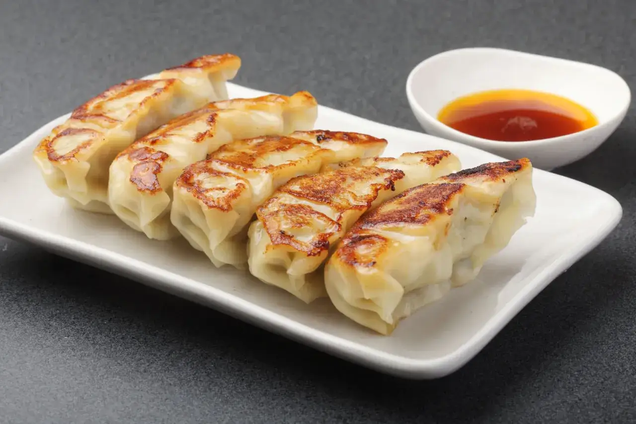 Złociste pierogi gyoza, chrupiące od spodu, podane na białym talerzu z miseczką sosu.