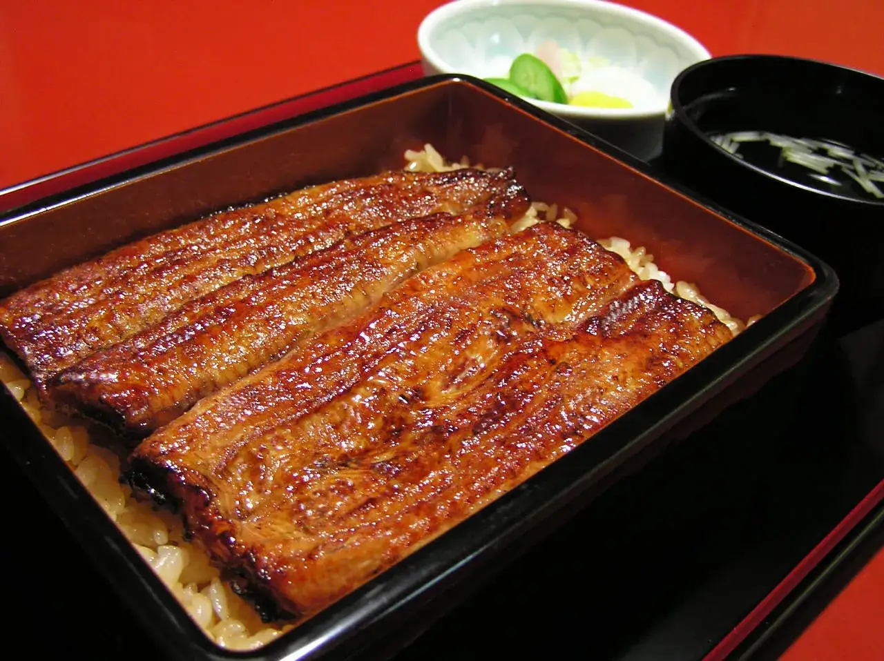 Lśniące kawałki unagi na ryżu, podane w drewnianym pudełku, obok miseczki z piklami i zupy.