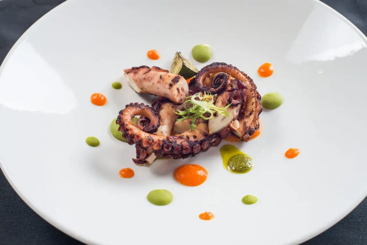 Grillowane macki ośmiornicy, podane z zielonym puree i pomarańczowym sosem, tworzą elegancką kompozycję na białym talerzu.