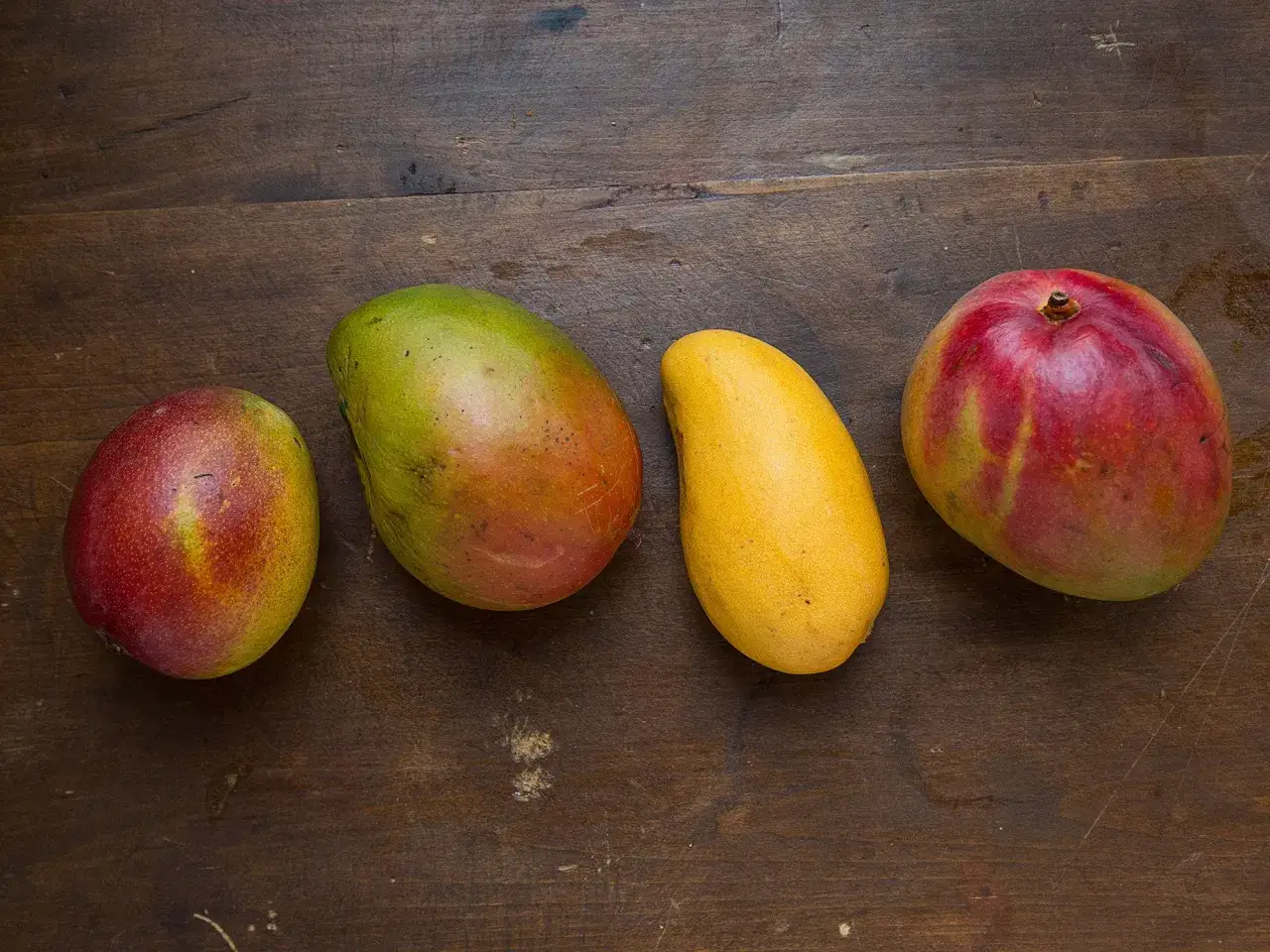 Cztery dojrzałe mango, o różnych odcieniach czerwieni, zieleni i żółci, leżą na drewnianym blacie.