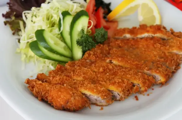 Chrupiący schab po japońsku, pokrojony w plastry, podany z sałatką z kapusty, ogórkiem, pomidorem i cytryną.