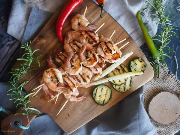 Grillowane krewetki po angielsku na szpadkach, podane z grillowaną cukinią, szparagami i papryczkami chili na drewnianej desce.
