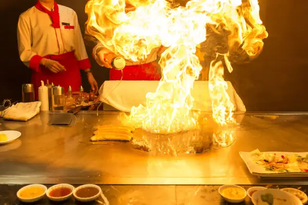 Szef kuchni tworzy spektakularny pokaz ognia podczas przygotowywania yakiniku. Płomienie unoszą się nad płytą grillową, a obok leżą kawałki mięsa.